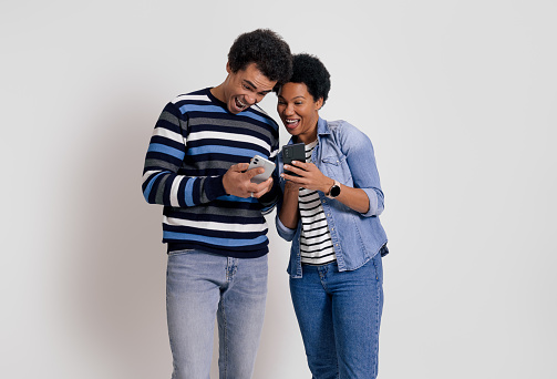 Ecstatic boyfriend and girlfriend showing mobile apps to each other and laughing on white background