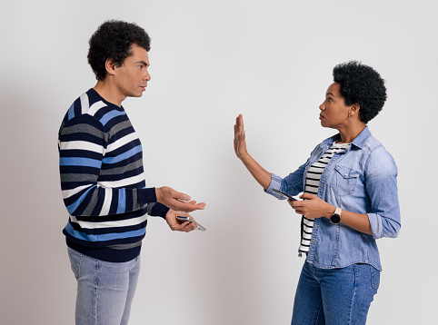 Angry young girlfriend using mobile phone and showing stop sign to boyfriend over white background