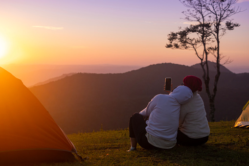 Couple is sitting by their tent during overnight camping and taking selfie while looking at the beautiful scenic sunset over the mountain for outdoor adventure vacation travel