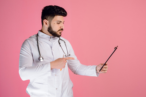 Side view of brunette doctor, intern holding folder, looking, pointing by finger, posing indoors. Isolated on pink studio background.