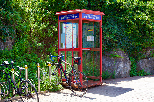 Donghae City, South Korea - July 29th, 2019: Bicycles rest beside the unique Chuam Chotdaebawi Certification Center, marked by a distinctive red phone booth-like structure without a phone, but with a stamp for cyclists to mark their bicycle certification passports. Set against a backdrop of a green and rocky hillside, this spot is part of South Korea's extensive bike path network featuring stamp booths across the country.