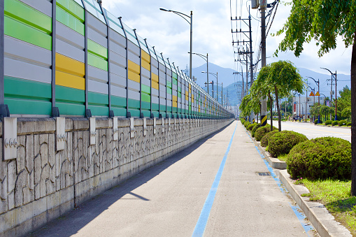 Donghae City, South Korea - July 29th, 2019: A scenic bike path runs parallel to a fifteen-meter-tall wall bordering Donghae Port, with majestic mountains in the backdrop and a road accompanied by a grassy median lying adjacent.
