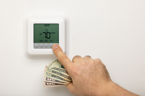 A hand holding money adjusting a thermostat on a wall.