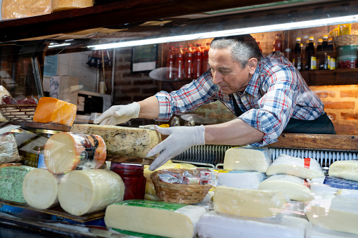 Senior man working at his small business organizing the cheese display at the deli - People at work concepts
