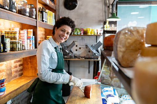 Latin American saleswoman cutting a block of cheese at a deli while smiling at camera - People at work concepts