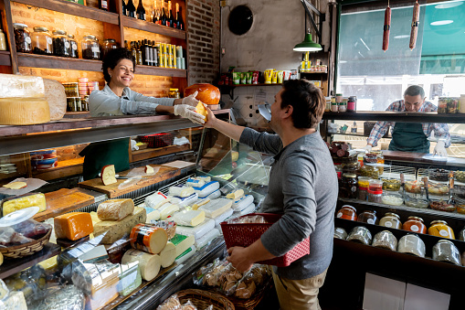 Friendly saleswoman handing a block of cheese to male customer buying products at the charcuterie - People at work concepts and consumerism concepts - Incidental worker at background