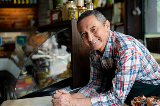 Senior man owner of a deli leaning on counter while smiling at the camera - Small business concepts