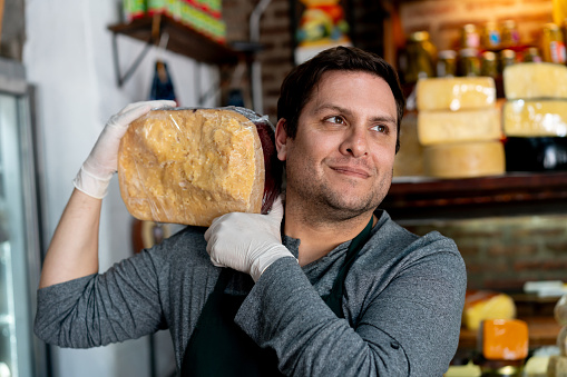 Male owner of a delicatessen carrying a cheese wheel on shoulder while looking away smiling - People at work concepts
