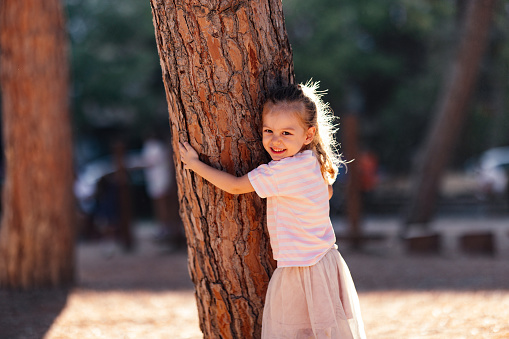 Smiling little girl looking at camera while embracing tree outdoors.