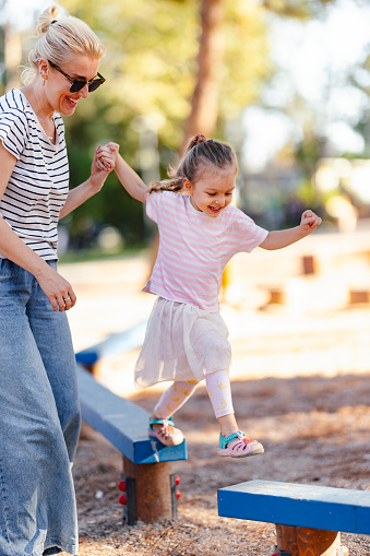 Happy little girl jumping on a blue wooden bench outdoors with the assistance of her mom.