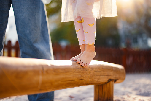 Close up photo of a barefoot girl legs walking on a wooden bench outdoors while her unrecognizable mother watching her.