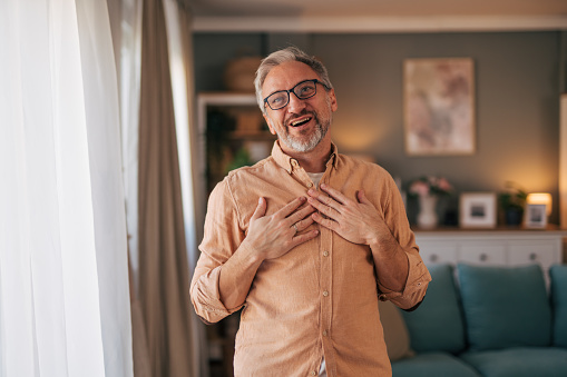 Mature man standing in his living room and gesturing like he is being complimented. He is looking happy.