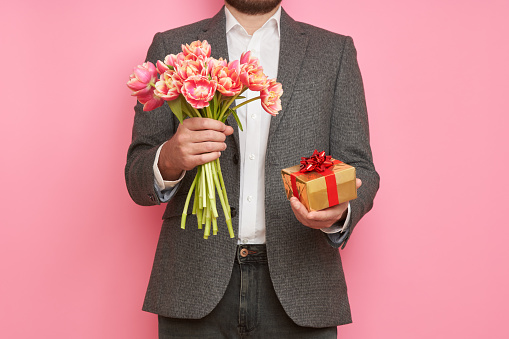 Romantic man in suit, with gift box and bouquet of tulips on isolated pink background, Valentine's Day, March 8