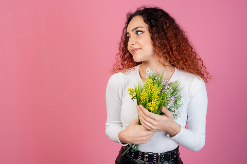 Front view of cheerful, young lady with red, curly hair posing indoors, holding flowers, smiling. Isolated on pink studio background.