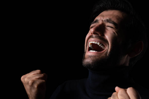 Mid-adult Caucasian male displaying intense anger. Wearing a white shirt, he clenches his fists, conveying powerful emotion against a dark background. The intense expression highlights raw human emotion and strong feelings.