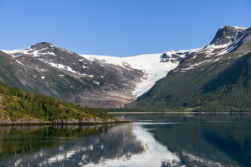 A serene fjord cradles the reflection of the majestic Svartisen Glacier, flanked by lush, green slopes under the bright Nordic sky
