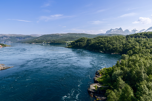 Vibrant greenery flanks the world-renowned Saltstraumen, where powerful water currents dance between Norwegian fjords under a blue sky