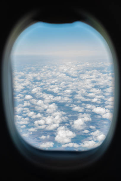 Clear view of fluffy clouds and blue sky framed by an airplane window. The image captures the timeless experience of flying above the earth’s surface.