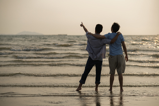 Two Young Asian men standing  on the beach, hug each other neck, point at the edge of sea with fun, cheerful, smile of happiness and hopefulness. Two men being with hope at beach concept at sunset time.