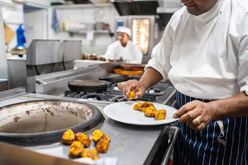 An unrecognisable person plating hot spicy chicken onto a clean white plate next to a tandoori oven in an industrial kitchen, with another worker busy behind