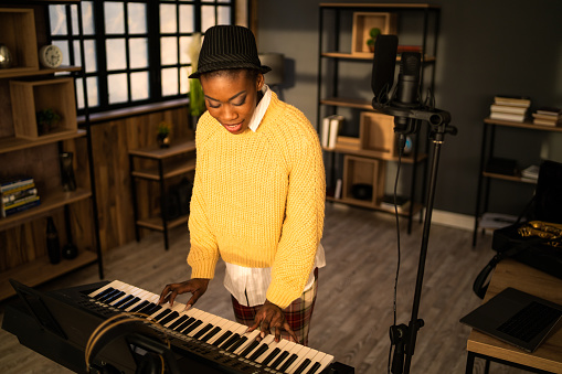 An African-American woman is rehearsing on a synthesizer in a dark modern music studio, wearing a bright yellow sweater and a black pork pie hat