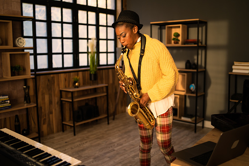 An African-American woman is rehearsing her music on her saxophone in a modern studio standing next to a keyboard