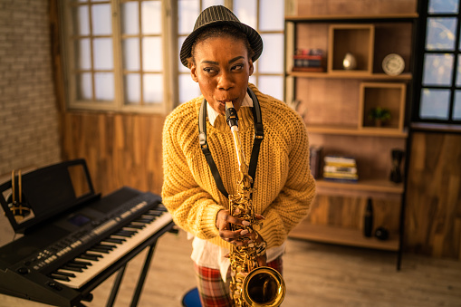 Front view of a woman with her lips around the mouthpiece of a saxophone concentrating on playing the instrument, she is rehearsing in a modern music studio