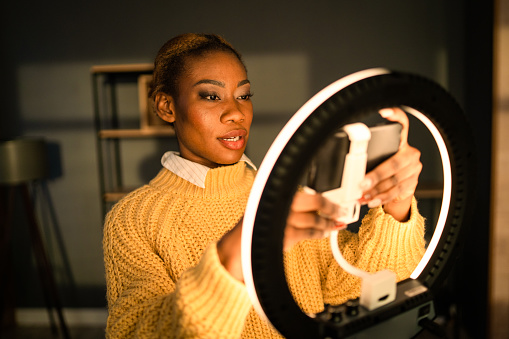 A woman wearing a bright yellow jumper is adjusting her cellphone in an LED stand, looking at her mobile screen, she is illuminated by the ring light