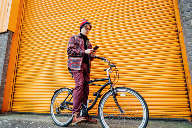 istock Young hipster man using mobile phone while sitting on cruiser bicycle near orange wall. Student using 5g network,rests after riding. Eco friendly transport for carbon footprint. Sustainable lifestyle. 2020458740