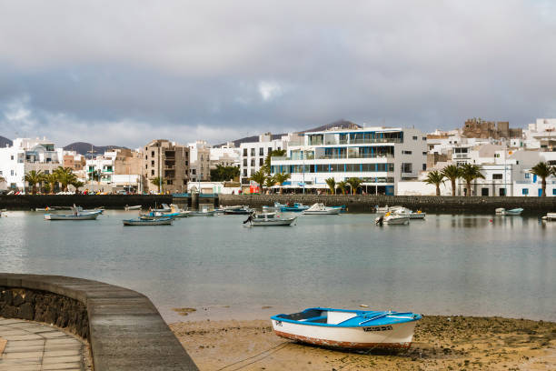 Arrecife, Lanzarote, Canary Islands, Spain - February 26th 2025:  The image depicts a serene coastal town with several small fishing boats anchored in the calm waters near the shore. The town features white buildings with blue accents, palm trees, and a rocky shoreline. The clear blue sky and tranquil sea create a picturesque and peaceful scene, highlighting the charm and simplicity of coastal life.