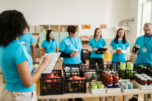 A female charity worker has a clipboard and a list to double check the groceries while the rest of the team have a coffee break in the background behind the tables full of donated food packed into crates ready for distribution