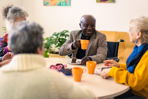 A small group of retired people are sitting around a table in a nursing home, drinking coffee from mugs and eating cake and biscuits