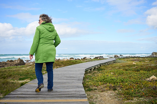 Rear view of a smiling mature woman walking along a wooden footpath by a rock beach on a windy afternoon