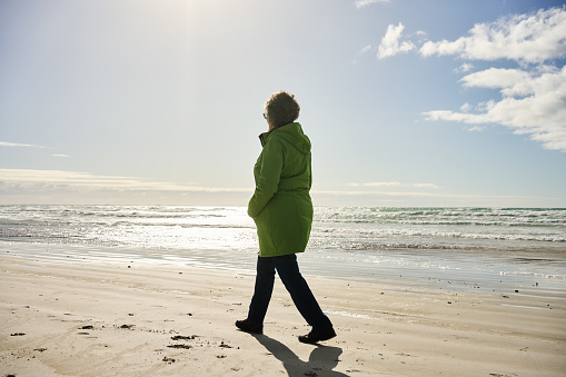 Mature woman looking at the view while walking alone along a sandy beach by the ocean on windy afternoon