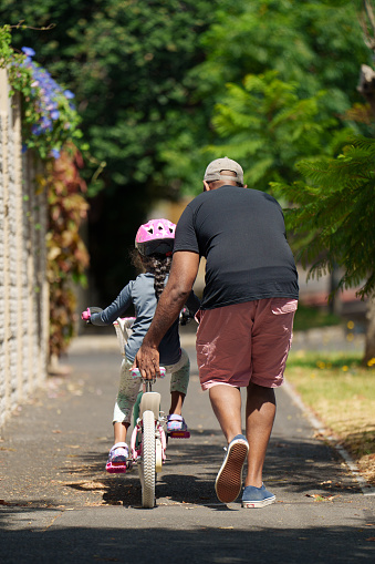 Rear view of a father teaching his little daughter how to ride a bicycle along a sidewalk outside in summer