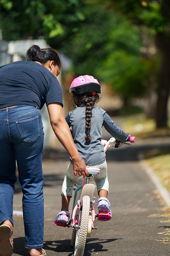 Rear view of a mother teaching her little daughter how to ride a bicycle along a sidewalk outside in summer