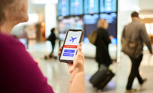 Over the shoulder view of a mature woman standing in an airport and receiving a flight cancellation notification on a mobile app on her phone