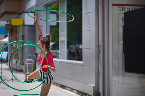 While standing on one leg, a street acrobat is spinning and twirling hoops on her arms and legs on a hot day in the city
