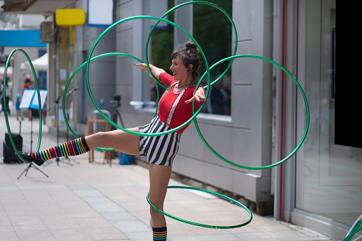 A street performer is twirling hoops around her arms and legs, while balancing on one leg, she is wearing a stripy colorful costume on a hot day in the city