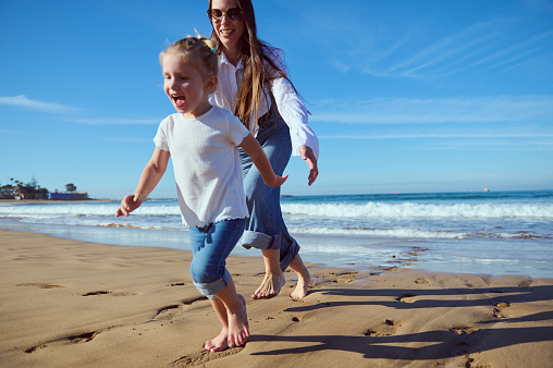 Full length portrait of a happy family of a Caucasian beautiful active little child girl and her mother laughing, smiling, playing together and running barefoot on the sandy beach