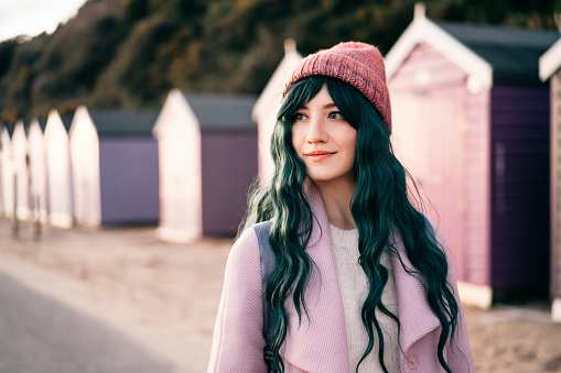 Stylish hipster woman with color hair in pink outfit and backpack walking along wooden beach huts on seaside. Off season Travel concept. Seasonal street fashion. Barbiecore style. Simple pleasures.