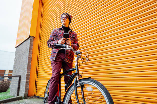 istock Young hipster man using mobile phone while sitting on cruiser bicycle near orange wall. Student using 5g network,rests after riding. Eco friendly transport for carbon footprint. Sustainable lifestyle 2020546677