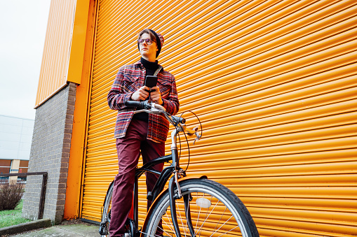 Young hipster man using mobile phone while sitting on cruiser bicycle near orange wall. Student using 5g network,rests after riding. Eco friendly transport for carbon footprint. Sustainable lifestyle.