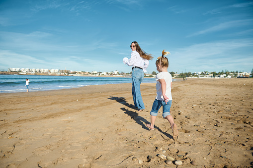 Rear view of a Caucasian happy mother and daughter smile while run along the Atlantic shore, enjoying beautiful waves washing their feet while pounding on the shore, leaving footsteps on the sand