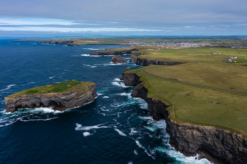 Aerial panoramic view of beautiful and green mountain of the famous tourist landmark Moher Cliff in Ireland with sea waves hitting rocks during day under cloudy sky.