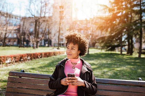 Young black woman enjoying in a beautiful spring time. She is sitting in a city park and using smart phone for messaging and communication.