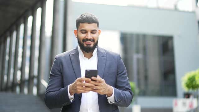 A smiling businessman is using a phone while walking along the street near an office building.