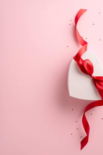 istock Perfect gift idea for her: vertical top view of heart-shaped present wrapped with red bow, surrounded by glitter on a soft pink backdrop, leaving space for your personalized message or advertisement 2020640630
