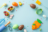 Easter-themed confectionery display. Top view of table adorned with baking toolsâwhisk, roller-pin, brush. Whimsical gingerbreads, chocolate eggs, candies, sprinkles on light blue background for text