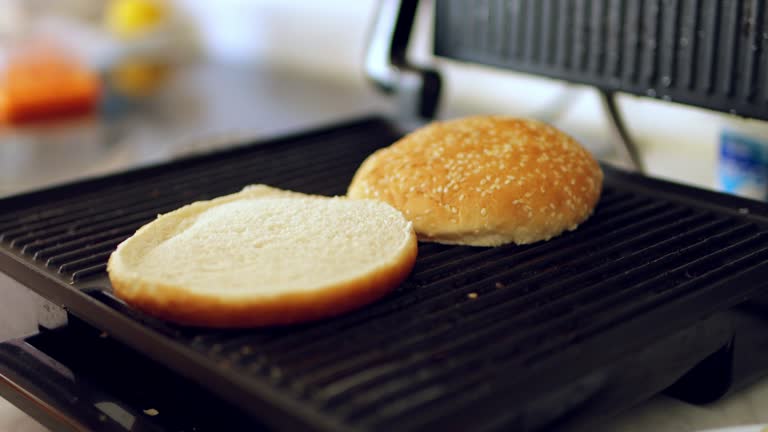 a man toasts a hamburger bun on an electric grill. There is a grill in the kitchen. The buns are ready